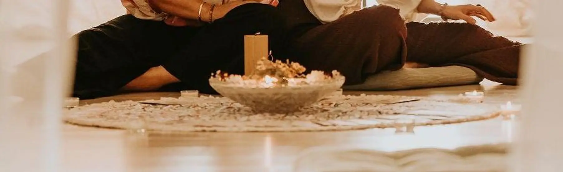 Au sol d’une salle d’atelier : un tapis clair, une coupe de fleurs séchées et une bougie, entourés de participantes assises.