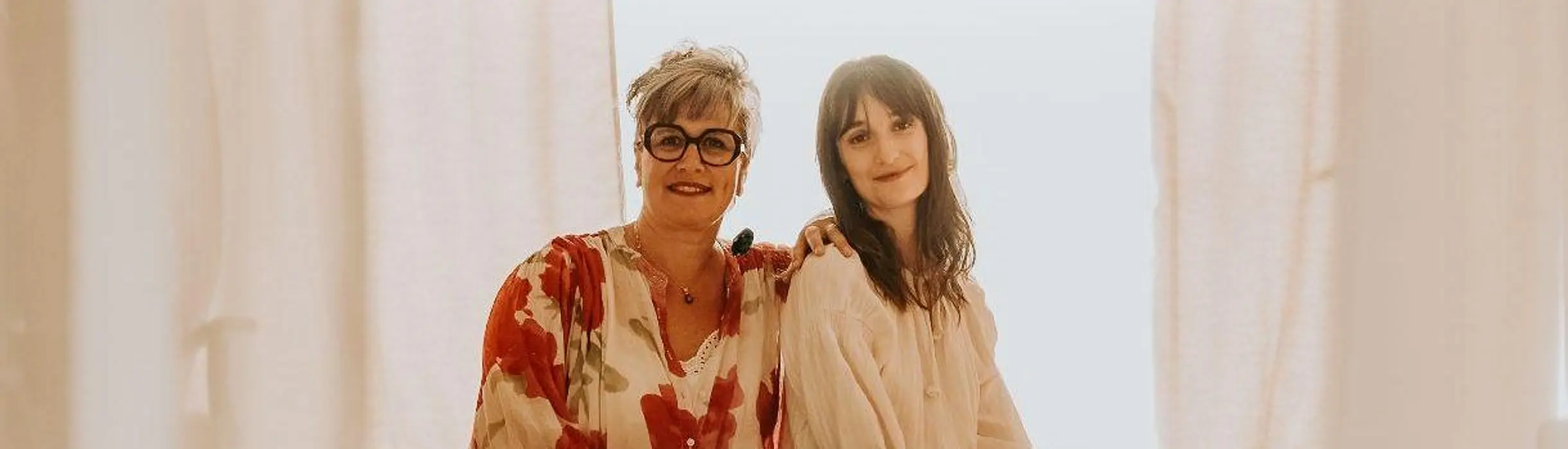Stéphanie et Morgane, debout côte à côte devant de grands voilages clairs, l’une la main posée sur l’épaule de l’autre.